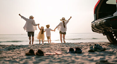 Beliebt bei Familien: mit dem eigenen Auto zum Badeurlaub an den Strand Rückansicht einer jungen vierköpfigen Familie in sommerlicher Kleidung, die an einem Sandstrand barfuß aufs Meer zuläuft. Im Vordergrund stehen ihre Schuhe neben dem geöffneten Familienauto im Sand.