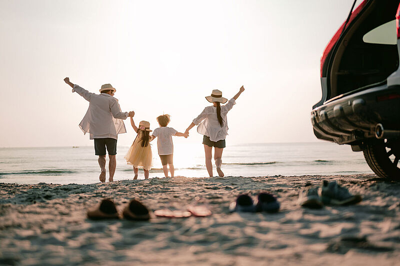 Rückansicht einer jungen vierköpfigen Familie in sommerlicher Kleidung, die an einem Sandstrand barfuß aufs Meer zuläuft. Im Vordergrund stehen ihre Schuhe neben dem geöffneten Familienauto im Sand.