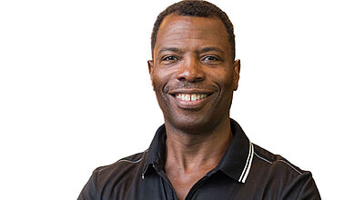 Porträt eines dunkelhäutigen, freundlich schauenden Mannes Mitte 30 mit kurzen, dunklen Haaren in einem schwarzen Poloshirt