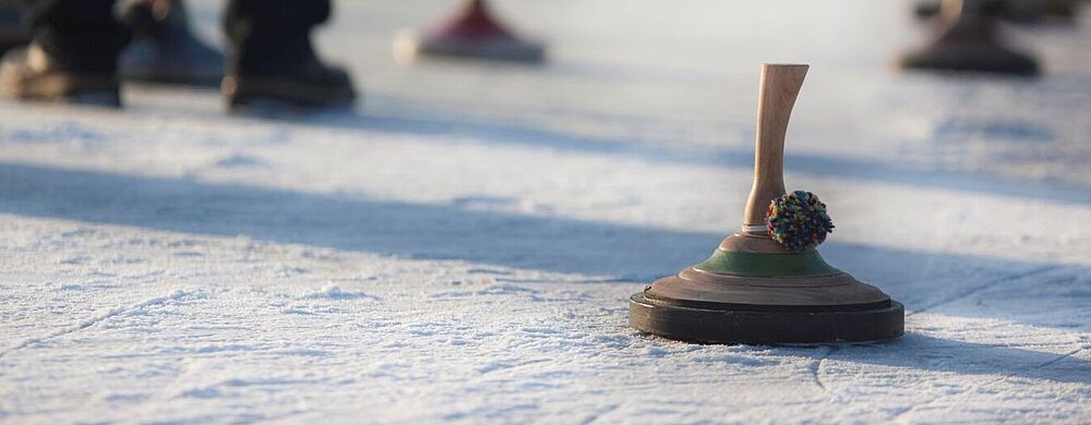Personen stehen auf einer vereisten Fläche und spielen Eisstockschießen; mehrere Eisstöcke liegen auf dem Eis, einer im Vordergrund ist scharf zu sehen.