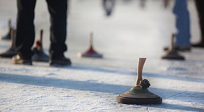 Personen stehen auf einer vereisten Fläche und spielen Eisstockschießen; mehrere Eisstöcke liegen auf dem Eis, einer im Vordergrund ist scharf zu sehen.