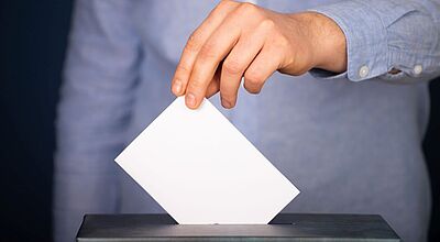 Nahaufnahme einer Hand, die einen weißen Wahlzettel in eine Wahlurne einwirft. Die Person trägt ein hellblaues Hemd.