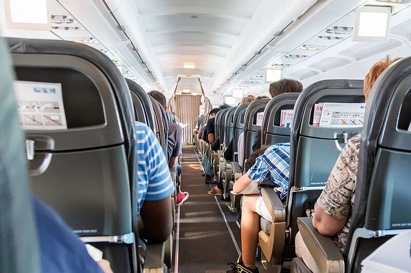 Blick in den Innenraum eines Flugzeugs. Auf den Plätzen sitzen Menschen. Ein Mann lehnt auf seinem Tisch. 