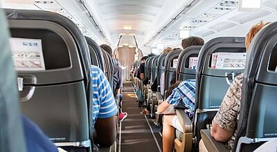 Blick in den Innenraum eines Flugzeugs. Auf den Plätzen sitzen Menschen. Ein Mann lehnt auf seinem Tisch. 