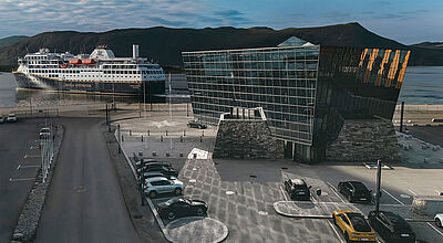 Der Havila-Hauptsitz in Fosnavag an Norwegens Westküste Futuristischer Firmensitz einer Kreuzfahrtreederei an der norwegischen Küste. Hinter dem gläsernen Gebäude passiert ein Schiff des Unternehmens – mit dunkelblauem Rumpf – die Zentrale. Im Hintergrund sind Berge zu sehen.