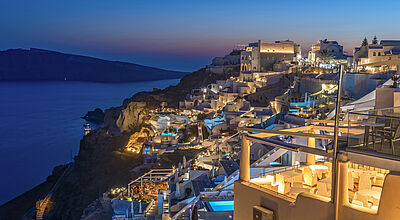 Weiße Häuser im Küstenstädtchen Oia auf der Insel Santorin an einem steilen Berghang, dazwischen blau beleuchtete Pools, blaue Stundezwischen 