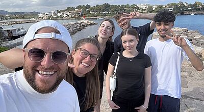 Eine Gruppe von sechs jungen Erwachsenen steht lachend auf einer Steinpromenade am Wasser. Im Hintergrund sind ein Hafen, Boote, ein Leuchtturm sowie weiße Gebäude und Bäume zu sehen. Die Personen posieren fröhlich für ein Selfie, machen Friedenszeichen und genießen offensichtlich einen sonnigen Tag am Meer.