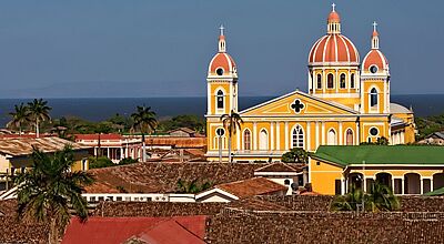 Sehenswert: die Kolonialstadt Granada in Nicaragua. Foto: FVA Nicaragua Panoramaansicht der kolonialen Stadt Granada in Nicaragua mit der markanten, gelb-roten Kathedrale von Granada im Zentrum. Die Kuppeln und Türme der Kathedrale ragen über die traditionellen Ziegeldächer der umliegenden Häuser hinweg. Im Hintergrund sind Palmen, grünes Landschaftsgebiet und der Nicaragua-See sichtbar. Die Architektur spiegelt den kolonialen Charme und die historische Bedeutung der Stadt wider.