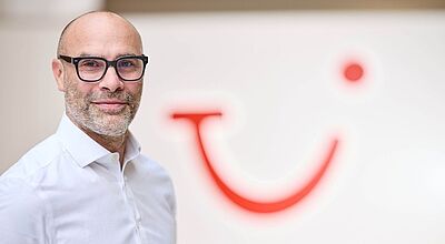 Ein Mann mittleren Alters mit Brille, kurzem Bart und weißem Hemd steht vor einer Wand mit dem roten Logo des Reisekonzerns TUI, das wie ein stilisiertes lachendes Gesicht aussieht. Er blickt lächelnd in die Kamera. Die Atmosphäre wirkt professionell und freundlich.