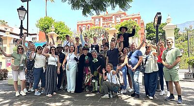 Eine fröhliche Gruppe von etwa 30 Personen steht im Freien vor einer historischen Kulisse mit bunten Gebäuden, Palmen und dekorativem Tor. Die Menschen lachen, winken und posieren für das Gruppenfoto. Zwei Personen in der Mitte sind kostümiert, eine davon trägt einen schwarzen Hut und lange schwarze Kleidung, die andere ein farbenfrohes Outfit mit grünem Halstuch. Im Hintergrund ist ein rotes Gebäude mit weißem Stuck sichtbar, vermutlich in mediterranem Stil. Das Wetter ist sonnig und klar.