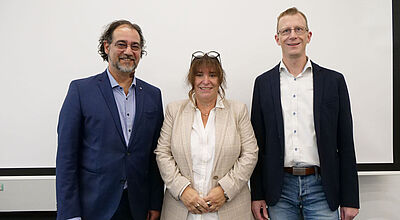 Zwei Männer und eine Frau im mittleren Alter stehen lächelnd in legerer Business-Kleidung vor einer beigen Wand