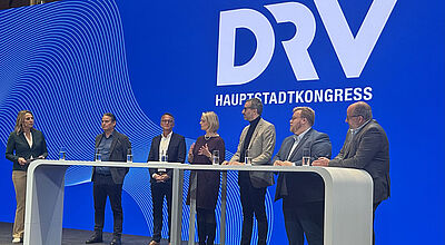 Podiumsdiskussion beim DRV Hauptstadtkongress: Sieben Teilnehmer stehen hinter weißem Pult vor blauem Hintergrund mit DRV-Logo.
