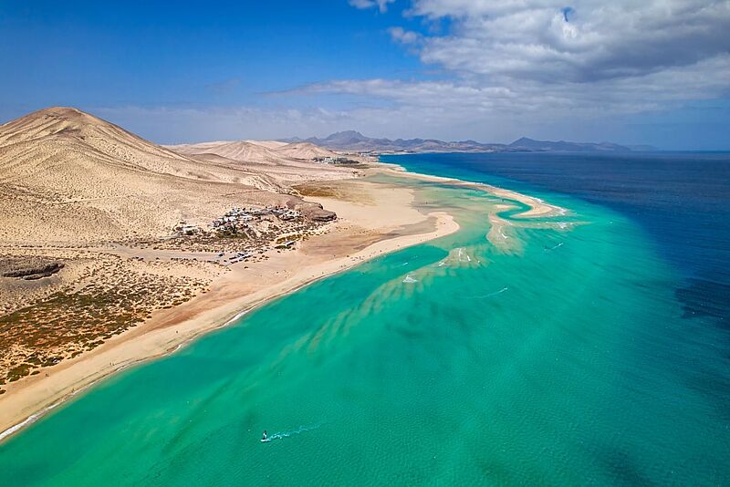 Luftaufnahme einer weitläufigen Sandlandschaft mit türkisblauem Meer. Eine schmale Sandbank ragt ins Wasser, dahinter erstrecken sich helle Dünen und karge Hügel unter blauem Himmel mit vereinzelten Wolken. Am Strand sind kleine Gebäude und geparkte Autos zu erkennen.