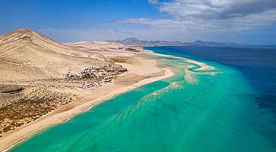 Fuerteventura (im Bild) und Lanzarote sind das Ziel einer Inforeise von Vtours auf die Kanaren Luftaufnahme einer weitläufigen Sandlandschaft mit türkisblauem Meer. Eine schmale Sandbank ragt ins Wasser, dahinter erstrecken sich helle Dünen und karge Hügel unter blauem Himmel mit vereinzelten Wolken. Am Strand sind kleine Gebäude und geparkte Autos zu erkennen.