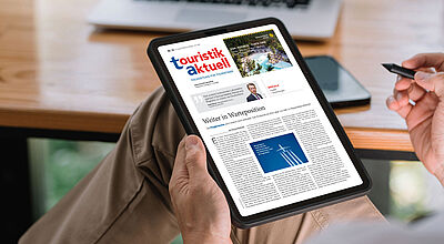 Eine Person sitzt entspannt mit übergeschlagenen Beinen und hält ein Tablet in der Hand, auf dem die digitale Ausgabe der Fachzeitschrift „touristik aktuell“ geöffnet ist. Die Titelseite zeigt unter anderem einen Artikel mit dem Titel „Weiter in Warteposition“ sowie eine Anzeige für die Dominikanische Republik. Im Hintergrund stehen ein Laptop und ein Smartphone auf einem Holztisch. Die Szene spielt sich in einer hellen, modernen Umgebung ab, vermutlich im Homeoffice oder Büro.