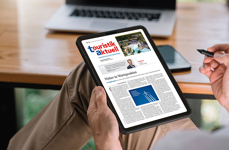 Eine Person sitzt entspannt mit übergeschlagenen Beinen und hält ein Tablet in der Hand, auf dem die digitale Ausgabe der Fachzeitschrift „touristik aktuell“ geöffnet ist. Die Titelseite zeigt unter anderem einen Artikel mit dem Titel „Weiter in Warteposition“ sowie eine Anzeige für die Dominikanische Republik. Im Hintergrund stehen ein Laptop und ein Smartphone auf einem Holztisch. Die Szene spielt sich in einer hellen, modernen Umgebung ab, vermutlich im Homeoffice oder Büro.
