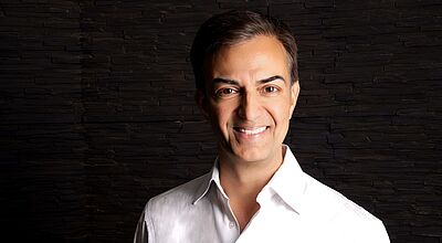 Andreas Höcherl ist bei Alltours jetzt für den Direktvertrieb und die Kundenbindung zuständig. Foto: Alltours Ein lächelnder Mann mit dunklem Haar trägt ein weißes Hemd und steht vor einer dunklen Wand aus horizontal geschichteten Steinen. Die Beleuchtung hebt sein Gesicht und die linke Seite seines Oberkörpers hervor.