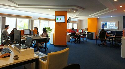 Modernes Büro mit blauem Teppich und orangefarbenen Akzenten, in dem mehrere Personen an Computern arbeiten. An der Wand ist ein leuchtendes Firmenschild mit der Aufschrift „gts Global Travel Services“ angebracht.