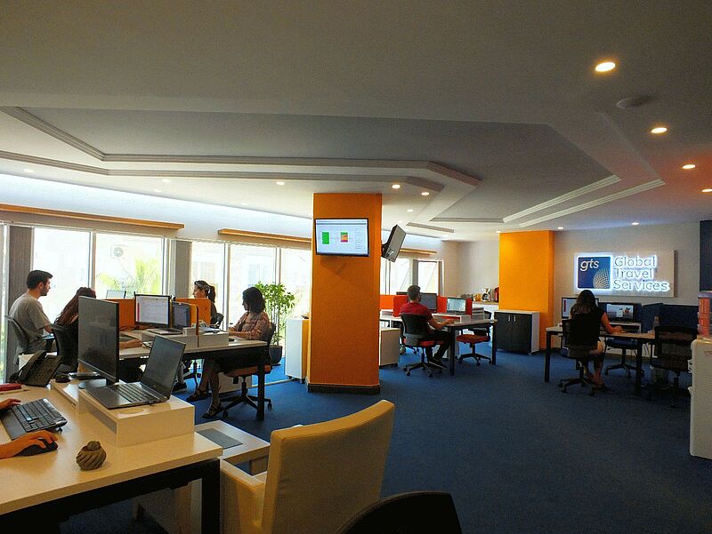 Modernes Büro mit blauem Teppich und orangefarbenen Akzenten, in dem mehrere Personen an Computern arbeiten. An der Wand ist ein leuchtendes Firmenschild mit der Aufschrift „gts Global Travel Services“ angebracht.