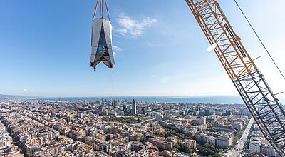 Ein großes Kreuzsegment wird mit einem Kran hoch über der Stadt Barcelona zur Sagrada Familia gehoben. Im Hintergrund sind die Stadt und das Mittelmeer zu sehen.
