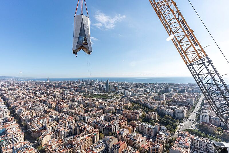 Die finale Bauphase der Sagrada Familia in Barcelona hat begonnen Ein großes Kreuzsegment wird mit einem Kran hoch über der Stadt Barcelona zur Sagrada Familia gehoben. Im Hintergrund sind die Stadt und das Mittelmeer zu sehen.