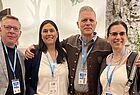 Vier Konferenzteilnehmer mit blauen Namensschildern posieren für ein Gruppenfoto, zwei tragen traditionelle Trachtenkleidung.