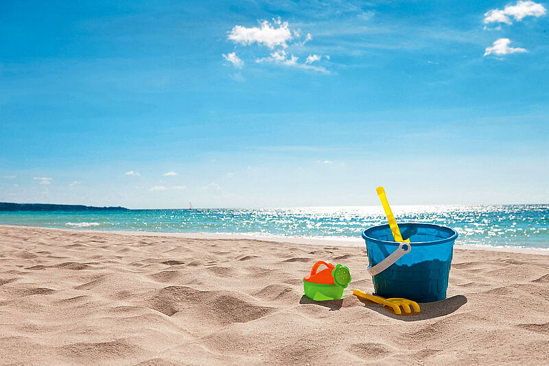 Ein sonniger Strand mit feinem Sand und türkisblauem Meer. Im Vordergrund stehen bunte Sandspielzeuge, darunter ein blauer Eimer mit gelbem Schaufelgriff, eine grüne Gießkanne und eine kleine Harke. Die Wellen glitzern in der Sonne, während am Horizont ein Segelboot zu sehen ist. Der Himmel ist blau mit wenigen weißen Wolken – eine idyllische Urlaubsszene