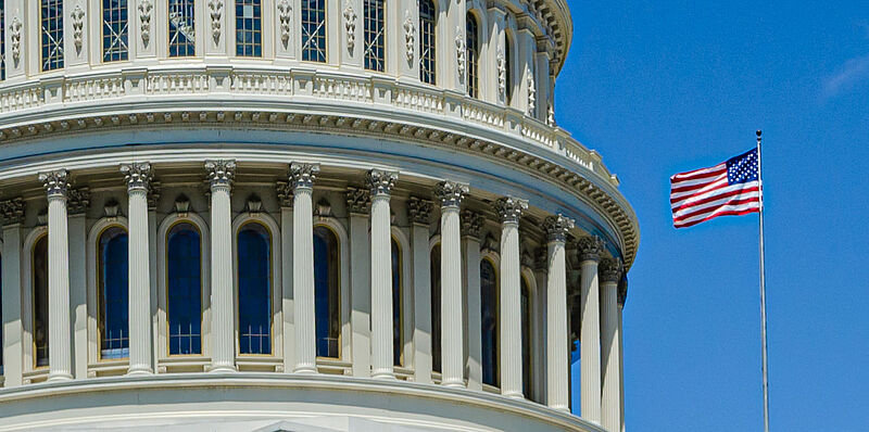 Detailaufnahme des Kapitols in Washington DC mit einer wehenden USA-Flagge vor blauem Himmel an einem sonnigen Tag