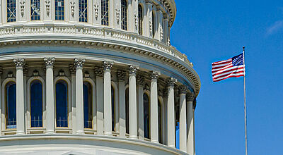 Detailaufnahme des Kapitols in Washington DC mit einer wehenden USA-Flagge vor blauem Himmel an einem sonnigen Tag