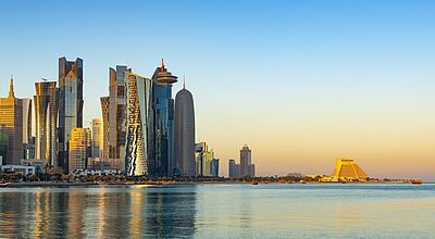 Moderne Skyline von Doha, Katar, bei Sonnenuntergang mit hoch aufragenden Wolkenkratzern, die sich im ruhigen Wasser der Bucht spiegeln; rechts im Bild das markante pyramidenförmige Sheraton Grand Doha Hotel.