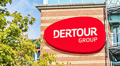 Außenansicht eines modernen Bürogebäudes mit rotem Dertour Group-Logo auf einer Backsteinfassade. Im Vordergrund sind grüne Bäume und ein blauer Himmel mit vereinzelten Wolken zu sehen.