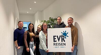Sechs Personen stehen nebeneinander in einem hellen Büroflur. Zwei von ihnen halten ein Schild mit dem Logo „EVR Reisen – Seit 1991“. Alle lächeln in die Kamera.
