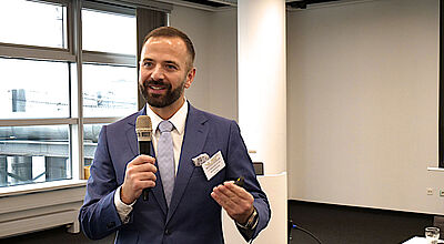Ein jüngerer Mann im grauen Anzug, weißem Hemd und Krawatte hält mit einem Mikrofon in der Hand einen Vortrag in einem Konferenzraum und gestikuliert