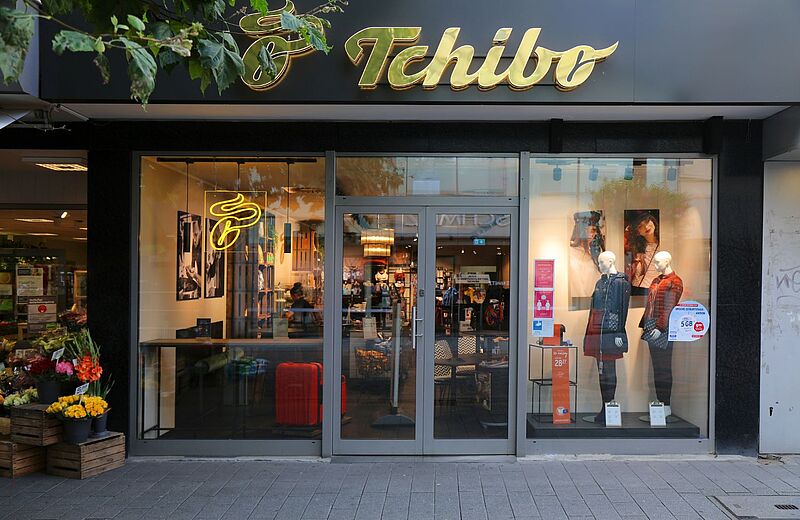 Eingang eines Tchibo-Geschäfts mit Glasfront, Schaufensterpuppen in herbstlicher Kleidung und leuchtendem gelbem Logo über der Tür; im Laden sind Regale und Produkte sichtbar, daneben ein Blumenstand auf dem Gehweg.