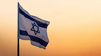 Für Israel gilt seit dem vergangenen Wochenende eine Reisewarnung. Foto: baona/iStock Israelische Flagge weht im Wind vor orangefarbenem Abendhimmel