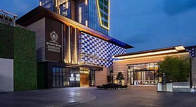 Das TUI Blue Sihanoukville ist nicht mehr als TUI Blue buchbar. Foto: TUI Modernes Hochhaus-Hotel bei Dämmerung, mit auffälliger Glasfassade, vertikalen Lichtelementen und begrünten Fassadenteilen. Der Eingang ist hell beleuchtet, der Platz davor leer und sauber gepflastert. Der Himmel ist klar und tiefblau.