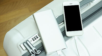 Ein weißes Handy liegt neben einer silberfarbenen Powerbank, die das Handy gerade lädt, auf einem weißen Rollkoffer.