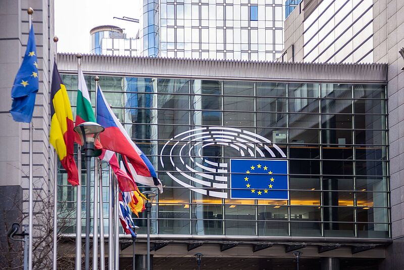 Fassade des Europäischen Parlaments in Brüssel mit dem EU-Logo und mehreren wehenden Flaggen der Mitgliedstaaten im Vordergrund.