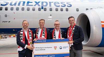 Vier Männer in Jacketts und Anzügen stehen mit Fanschals des VfB Stuttgart vor einem Flugzeug der Airline Sun Express. Sie alle halten einen großen symbolischen Boarding Pass, auf dem eine neue Partnerschaft der Airline und des Fußball-Bundesligisten verkündet wird.