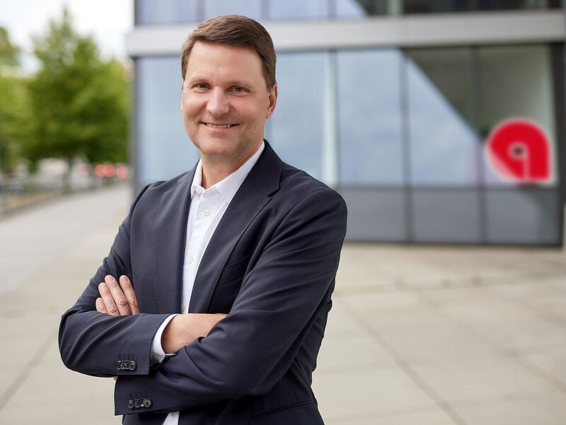 Mit dem neuen Partnermodell „Alltours Plus“ will Vertriebschef Georg Welbers weitere Anreize für den Verkauf von Alltours setzen. Foto: Alltours Porträt eines lächelnden Mannes mittleren Alters mit braunen Haaren im dunklen Anzug und vor der Brust verschränkten Armen. Er steht vor einem Gebäude mit Glasfassade.