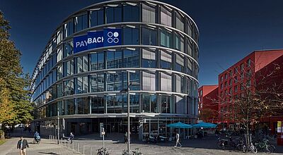  Modernes, rundes Bürogebäude mit Glasfassade und Payback-Logo an der oberen Ecke; im Vordergrund eine gepflasterte Fußgängerzone mit Fahrrädern, Fußgängern und einem Café mit blauen Sonnenschirmen; im Hintergrund rote Gebäude bei sonnigem Wetter.