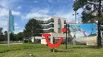 Außenaufnahme eines modernen Bürogebäudes mit Glas- und Betonfassade, davor eine große rote 3D-Skulptur des TUI-Logos in Form eines stilisierten lächelnden Gesichts. Links im Bild wehen zwei hellblaue Fahnen mit dem gleichen Logo. Im Hintergrund steht ein großes Werbeplakat, das einen sonnigen Strand mit Meer und das TUI-Logo zeigt.