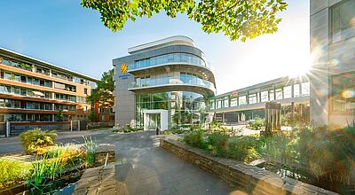 Moderne Gebäudefassade mit viel Glas und geschwungenen Balkonen, daneben ein begrünter Innenhof mit Wasserbecken und Steinmauern. Im Hintergrund verbindet eine verglaste Brücke zwei Gebäudeteile. Die Sonne scheint durch die Bäume und erzeugt Lichtreflexe.“