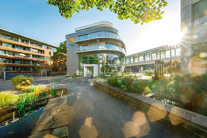 Moderne Gebäudefassade mit viel Glas und geschwungenen Balkonen, daneben ein begrünter Innenhof mit Wasserbecken und Steinmauern. Im Hintergrund verbindet eine verglaste Brücke zwei Gebäudeteile. Die Sonne scheint durch die Bäume und erzeugt Lichtreflexe.“