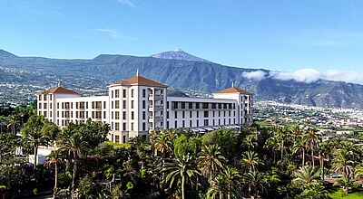 Hinter einem grünen Palmengarten erhebt sich ein ehrwürdiges weißes Gebäude mit quadratischem Grundriss und rotem Dach. Im Hintergrund ist der Berg Teide zu sehen. 