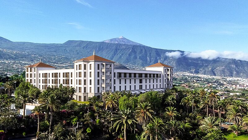 Hinter einem grünen Palmengarten erhebt sich ein ehrwürdiges weißes Gebäude mit quadratischem Grundriss und rotem Dach. Im Hintergrund ist der Berg Teide zu sehen. 