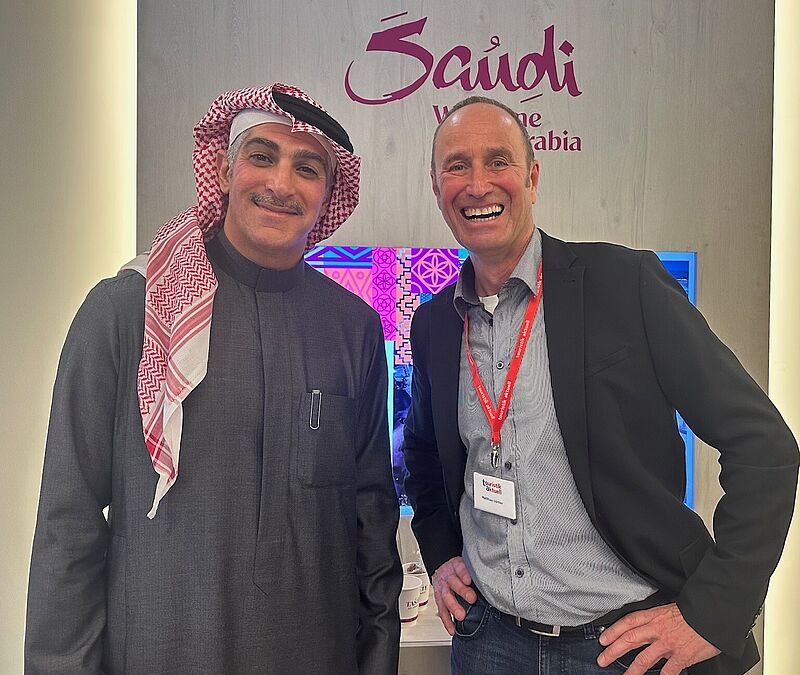 Zwei Männer stehen lächelnd vor einer Werbewand von Saudi-Arabien, einer in traditioneller arabischer Kleidung, der andere mit Hemd und Sakko 