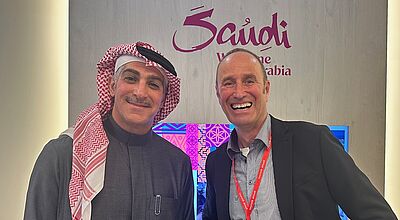 Zwei Männer stehen lächelnd vor einer Werbewand von Saudi-Arabien, einer in traditioneller arabischer Kleidung, der andere mit Hemd und Sakko 
