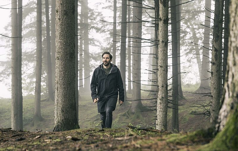Ein Mann mit dunklem Haar und Vollbart läuft durch einen Wald, zwischen den Stämmen der Fichten hängt leichter Nebel.