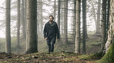 Ein Mann mit dunklem Haar und Vollbart läuft durch einen Wald, zwischen den Stämmen der Fichten hängt leichter Nebel.